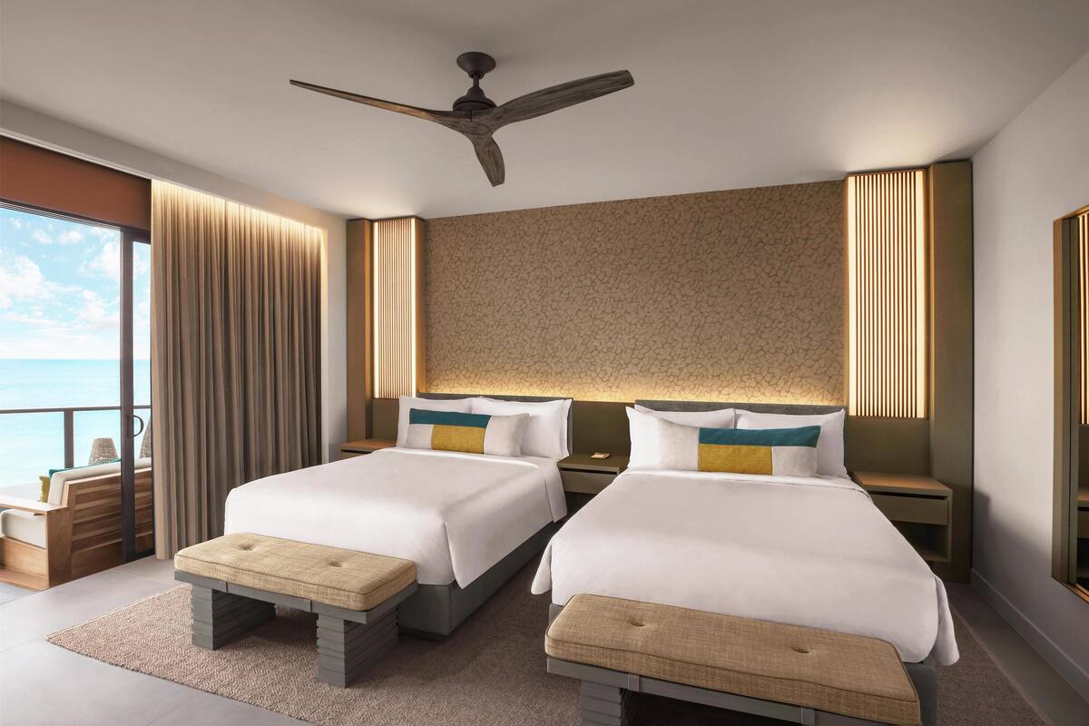 A modern hotel room with two beds, a ceiling fan, and a large window offering a view of the sea.
