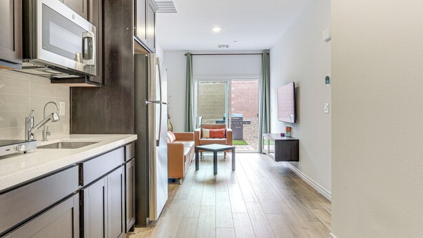 A modern kitchen with dark cabinets, a stainless steel microwave, and a large sink. There is a dining area with a table and chairs, and a TV mounted on the wall.