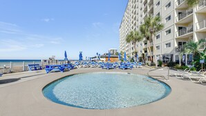 Une piscine entourée de chaises et de parasols bleus, avec un immeuble de grande hauteur en arrière-plan, doté de balcons et de palmiers.