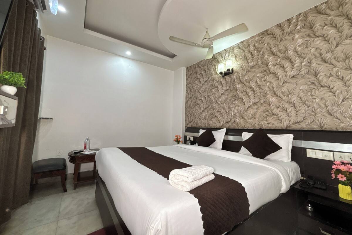 A hotel room with a large bed, a ceiling fan, a wall-mounted light fixture, and a small bedside table with a lamp.