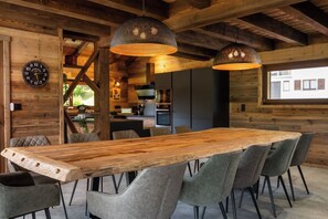 A wooden dining table with grey upholstered chairs, a large wall clock, and two hanging pendant lights in a rustic interior. - ✨ Chalet Marcel – Luxury Chalet with Hot Tub, Sauna & Forest Setting (Gérardmer)