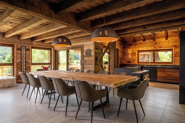 A spacious dining area with a long wooden table and modern chairs, a kitchen with wooden cabinets, and large windows offering a view of the outdoors.