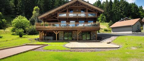 A two-story wooden chalet with a balcony, surrounded by a well-maintained lawn and a forested backdrop.