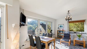A modern living room with a dining table, chairs, a bookshelf, a TV mounted on the wall, and a view of the outdoors. - Apartment 'Burgblick' with Balcony and Wi-Fi (Sankt Goar)