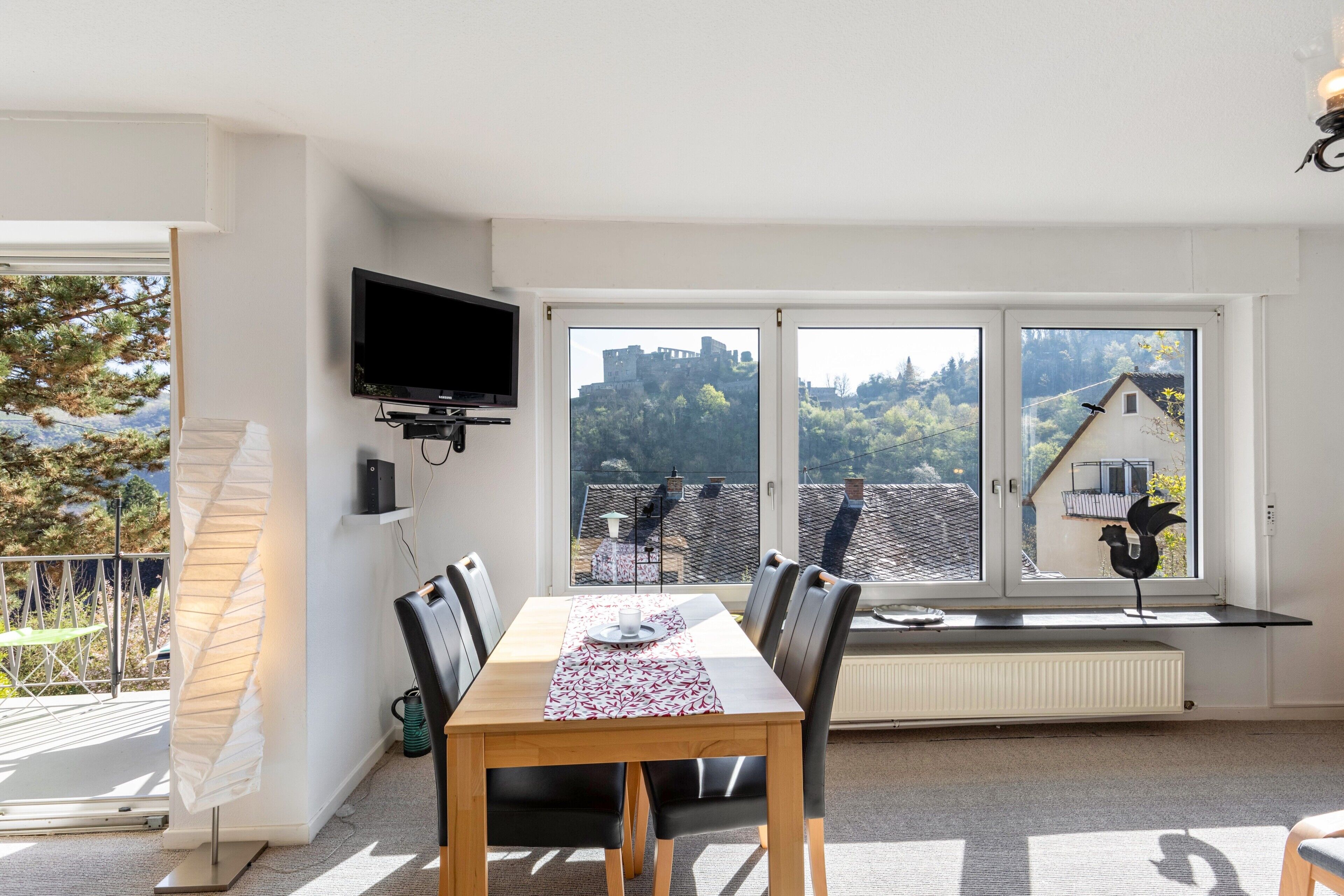 Ein Essbereich mit Tisch, Stühlen, einem Fernseher und großen Fenstern mit Blick auf ein Schloss und Bäume.
