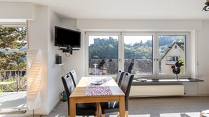 A dining area with a table, chairs, a TV, and large windows with a view of a castle and trees. - Apartment 'Burgblick' with Balcony and Wi-Fi (Sankt Goar)