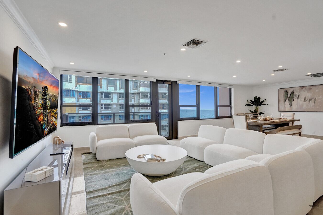 A modern living room with a large window, white sofas, a coffee table, and a flat-screen TV.