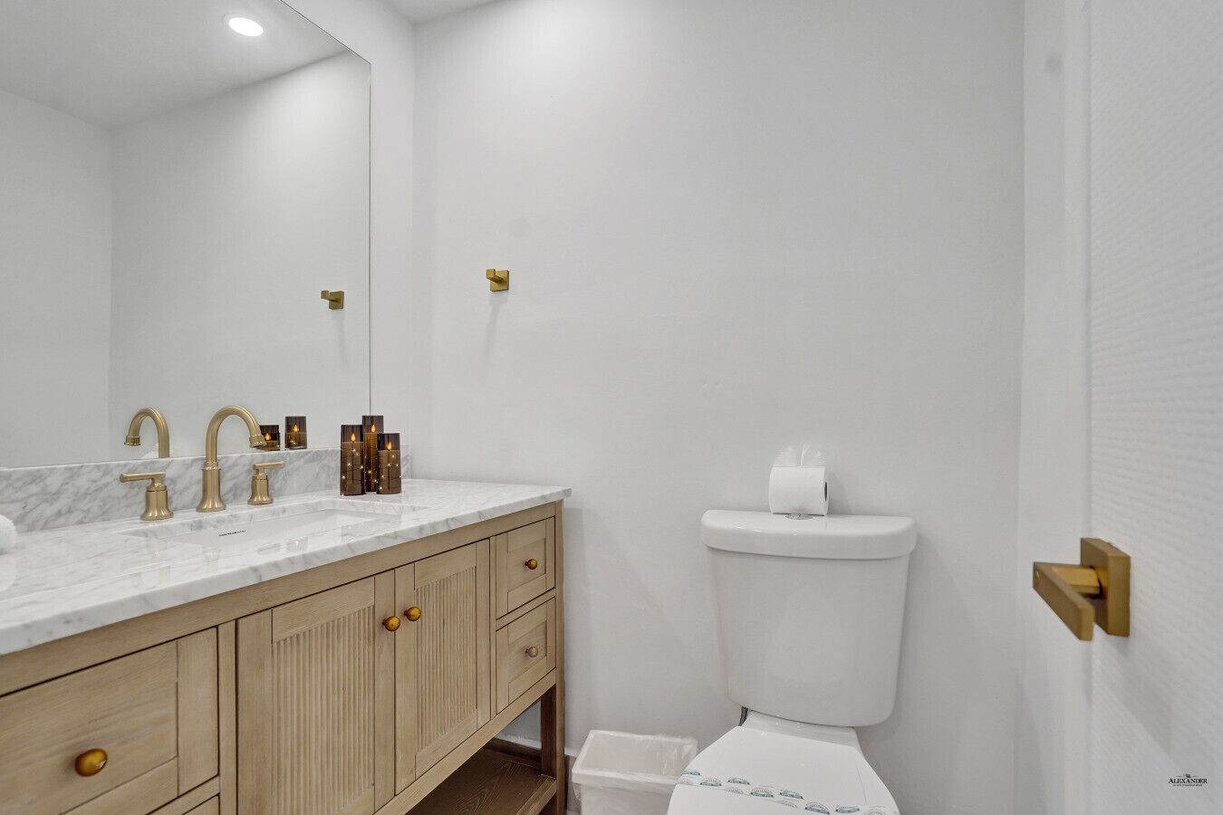 A bathroom with a marble countertop, gold faucets, a toilet, and a wooden vanity.