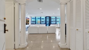 Un hall d’entrée spacieux, doté de colonnes blanches, d’un sol carrelé de couleur beige et offrant une vue sur un salon moderne avec un canapé et de grandes fenêtres.
