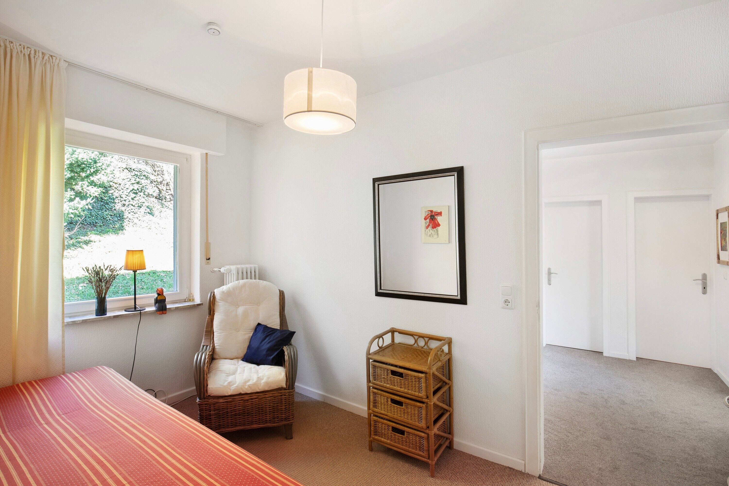 A room with a bed, a chair, a window with curtains, a wall-mounted lamp, a framed picture, and a door.