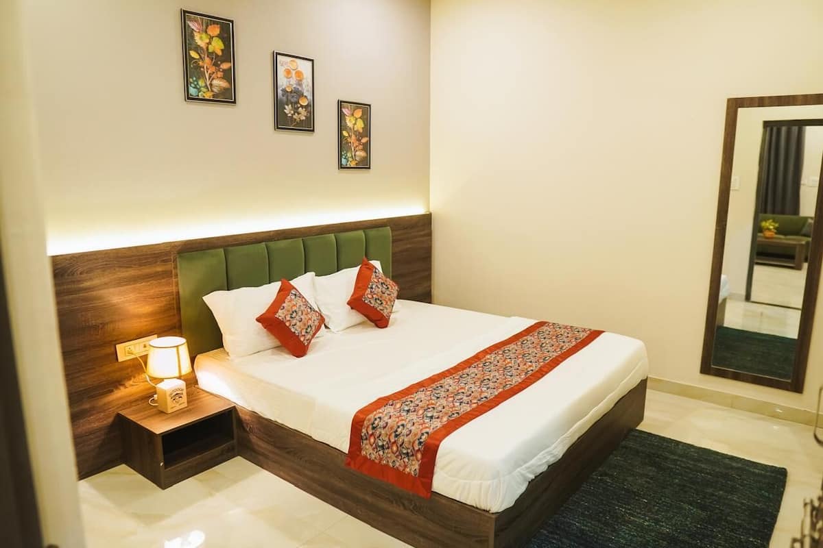 A neatly made bed with a green headboard, white bedding, and a patterned throw. There is a nightstand with a lamp and a small decorative item. Three framed floral artworks are hung on the wall above the bed.