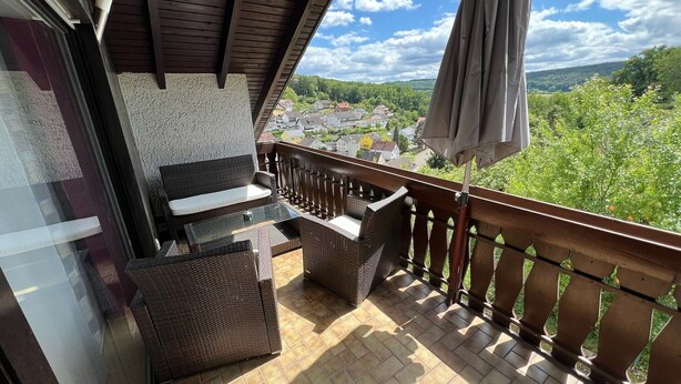 Un balcon agrémenté de meubles en osier, d’une table en verre et offrant une vue sur la ville et la verdure.