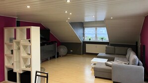 Un appartement moderne de style « loft », doté d’un plafond en pente, d’étagères intégrées, d’un canapé gris et d’un téléviseur.