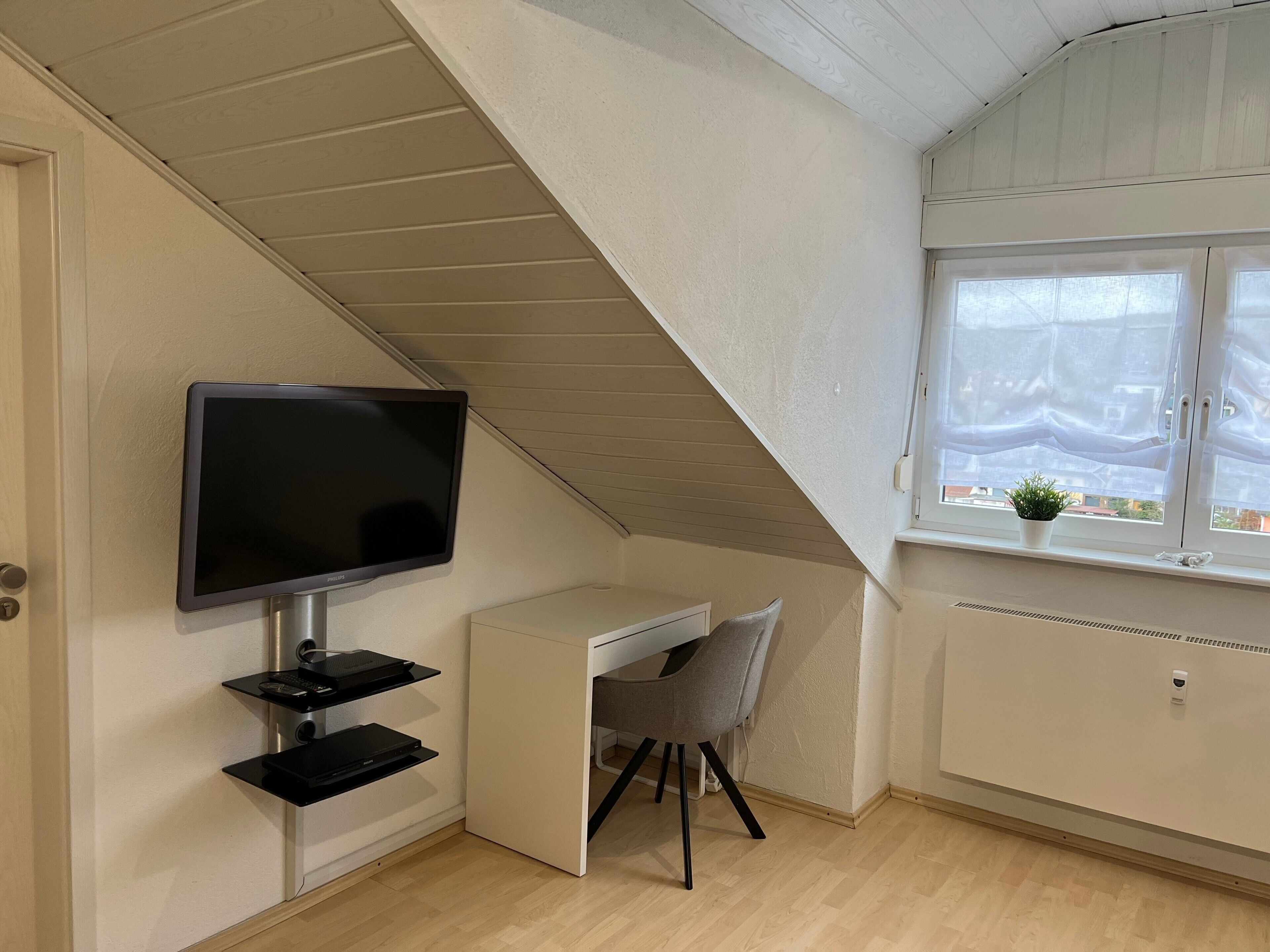 A room with a TV mounted on the wall, a desk with a chair, a window with a view, and a radiator.