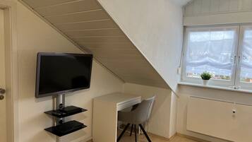 A room with a TV mounted on the wall, a desk with a chair, a window with a view, and a radiator.