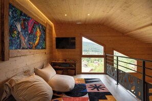 A cozy cabin interior with a wooden ceiling, a colorful abstract painting, a sofa, a flat-screen TV, and a view of the outdoors.