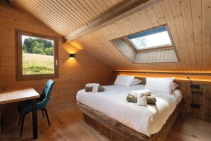 A bedroom with a wooden ceiling, a skylight, a bed with pillows and towels, a wooden desk, and a chair.