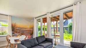 A modern living room with a grey sofa, a white coffee table, and a dining area with a white table and chairs. Large windows provide a view of the outdoors.