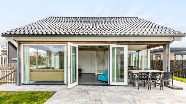 A modern house with a tiled roof, a patio with a dining table and chairs, and a glass door leading to a living area with a sofa and a blue chair. - Duinzicht Renesse by Interhome (Renesse)