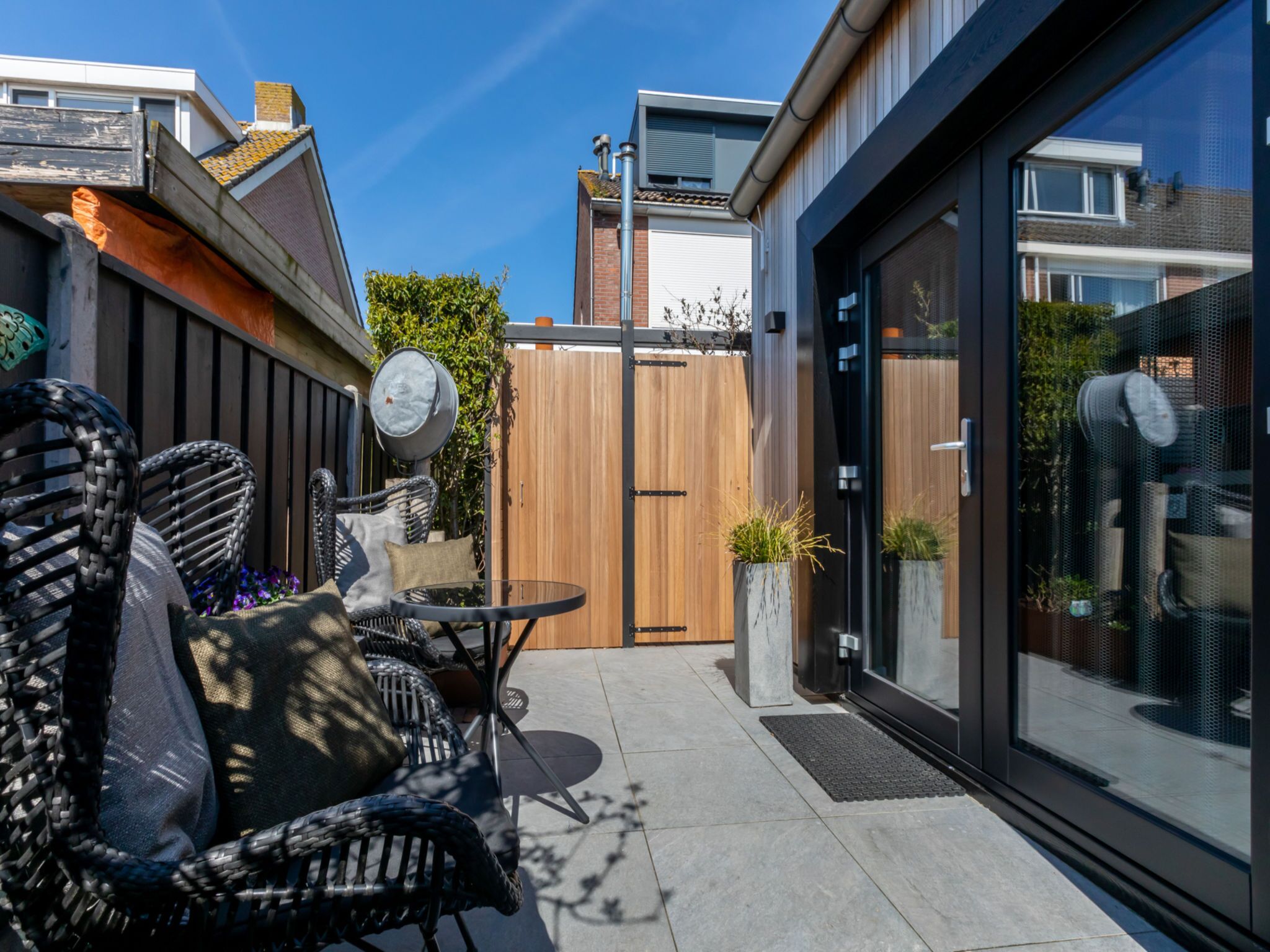 Une terrasse extérieure moderne, dotée de meubles en osier, d’une clôture en bois et d’une porte vitrée donnant sur un jardin.