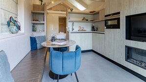 Uma cozinha moderna com forno embutido, área de jantar com mesa redonda e cadeiras azuis, e sala de estar com sofá e planta.