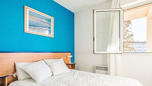 Ein Hotelzimmer mit Bett, Nachttisch, Nachttischlampe, einem Fenster mit Blick auf Gebäude und einem Bild an der Wand.