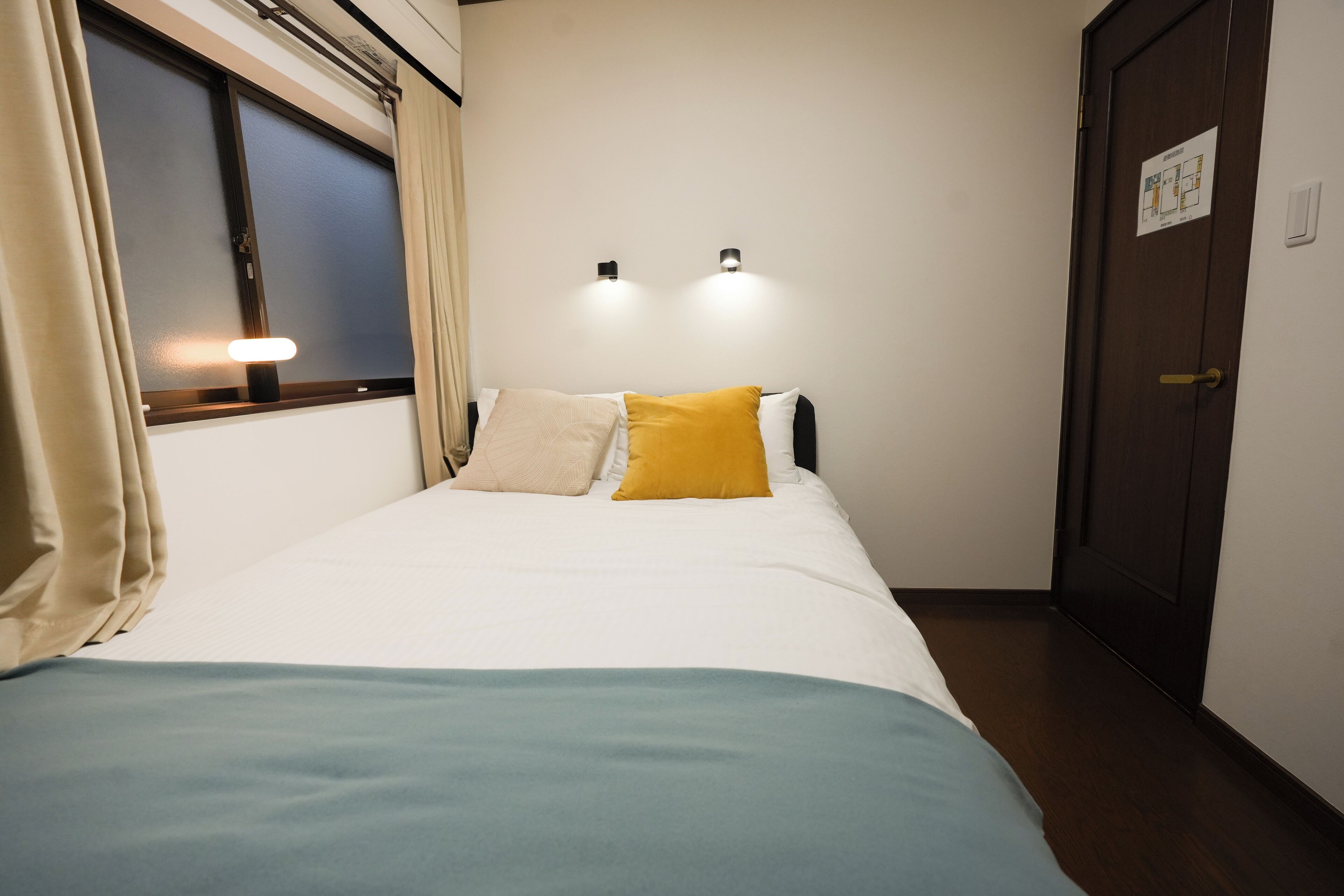 Hotelkamer met een bed, een raam met gordijnen, twee wandlampen en een deur met een bordje.