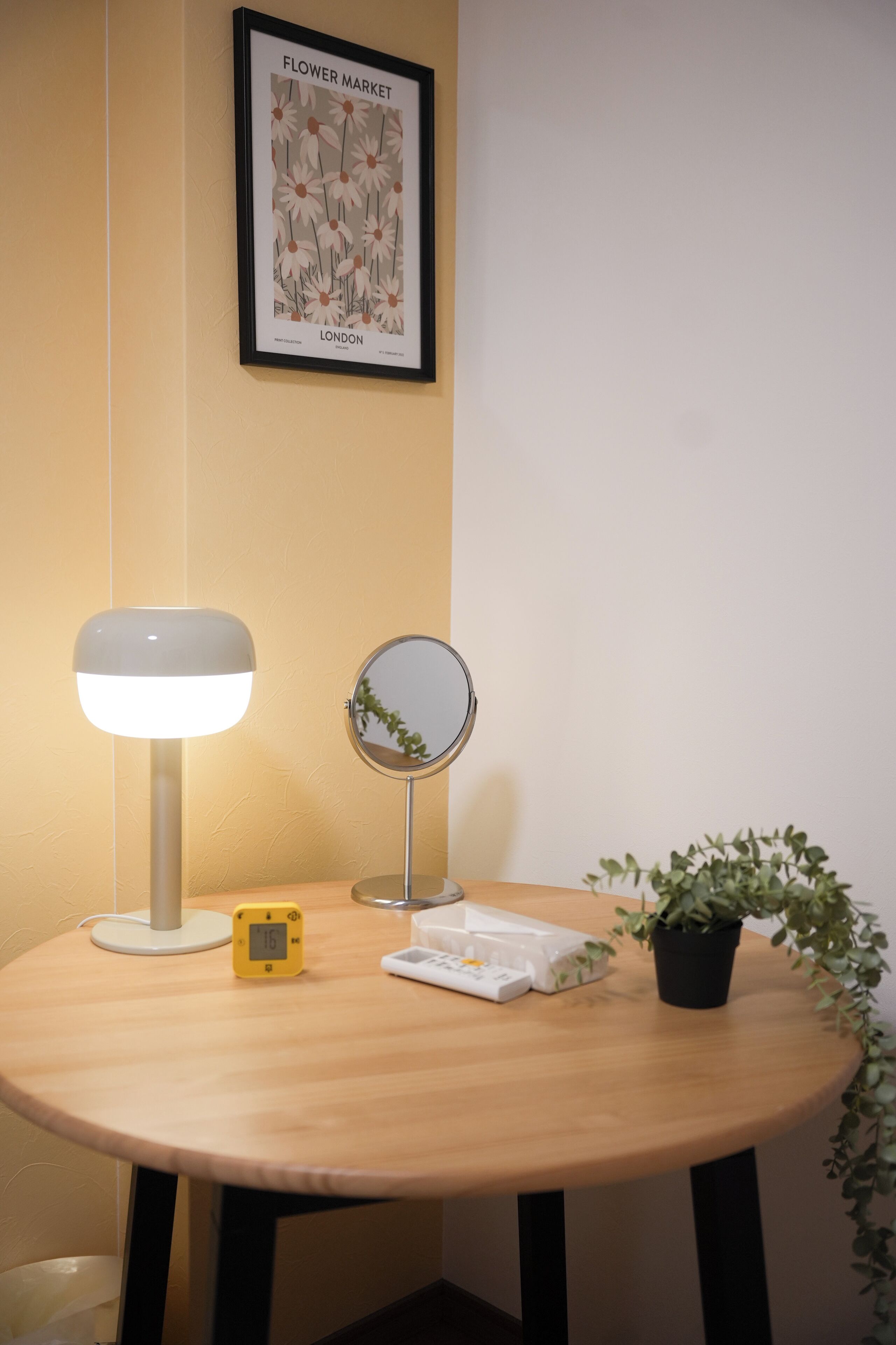 Una mesa redonda de madera con una lámpara de escritorio blanca, un espejo pequeño, un reloj digital y una planta en maceta.