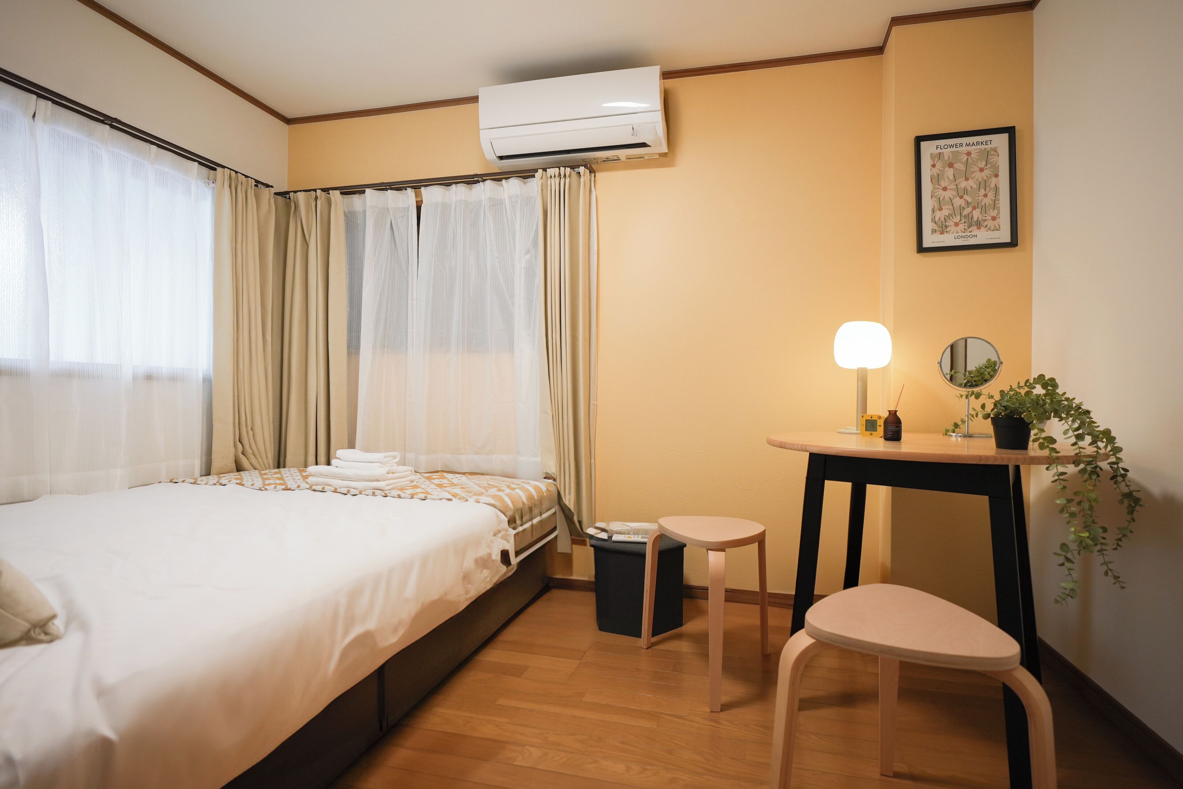 A hotel room with a bed, a small table with a lamp, a chair, a trash bin, and a potted plant.