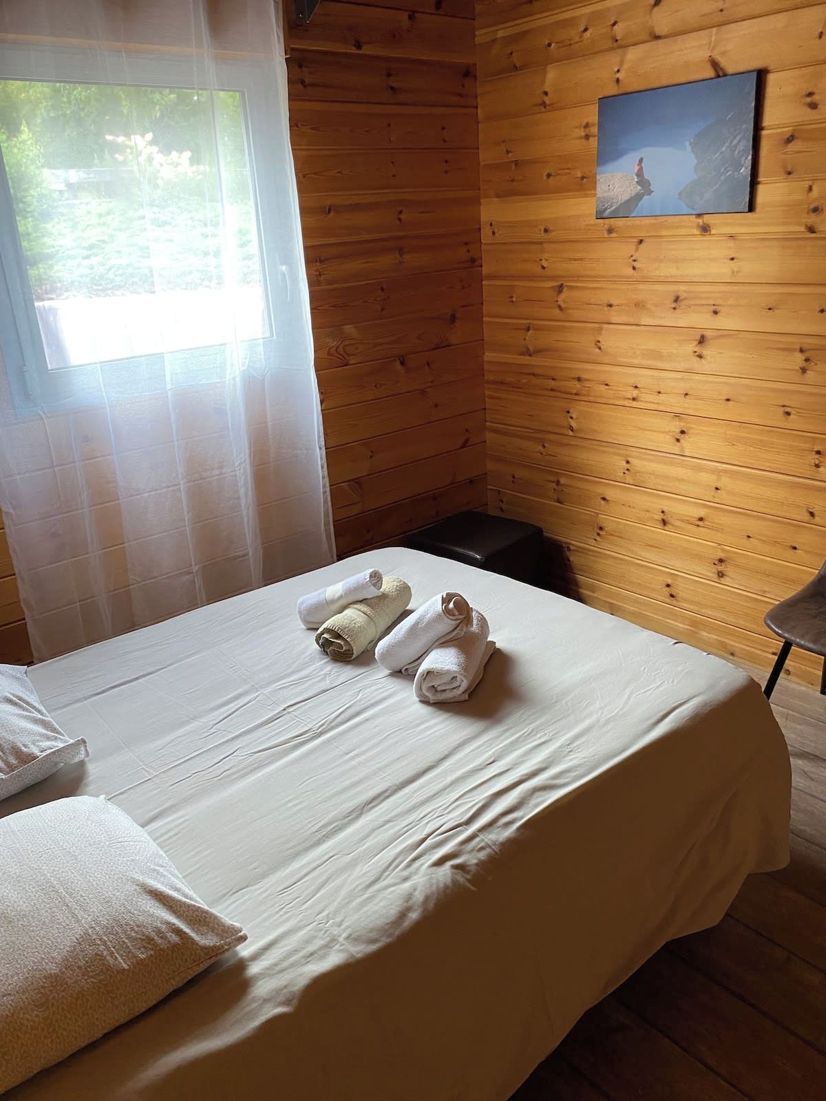 A wooden cabin room with a bed, towels, a window, and a painting on the wall.