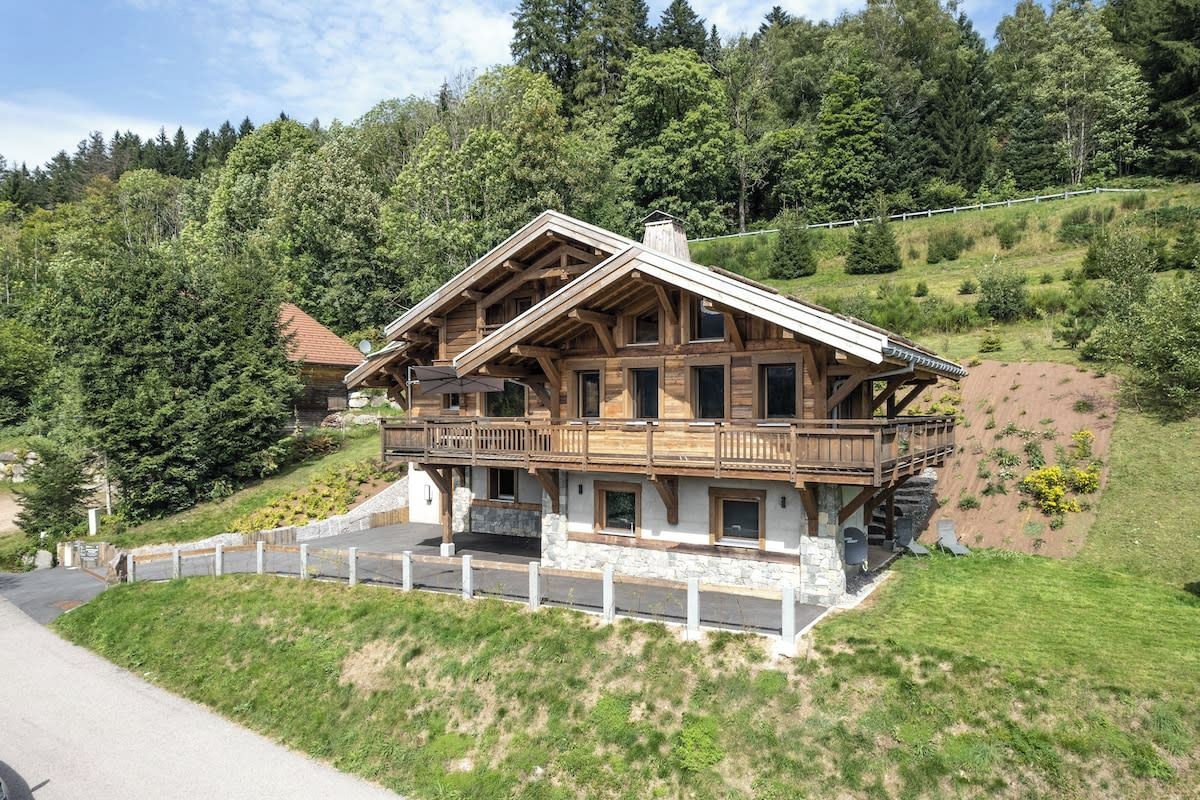 Un chalet en bois à deux étages, doté d’un balcon, situé sur une colline et offrant une vue sur une zone boisée.