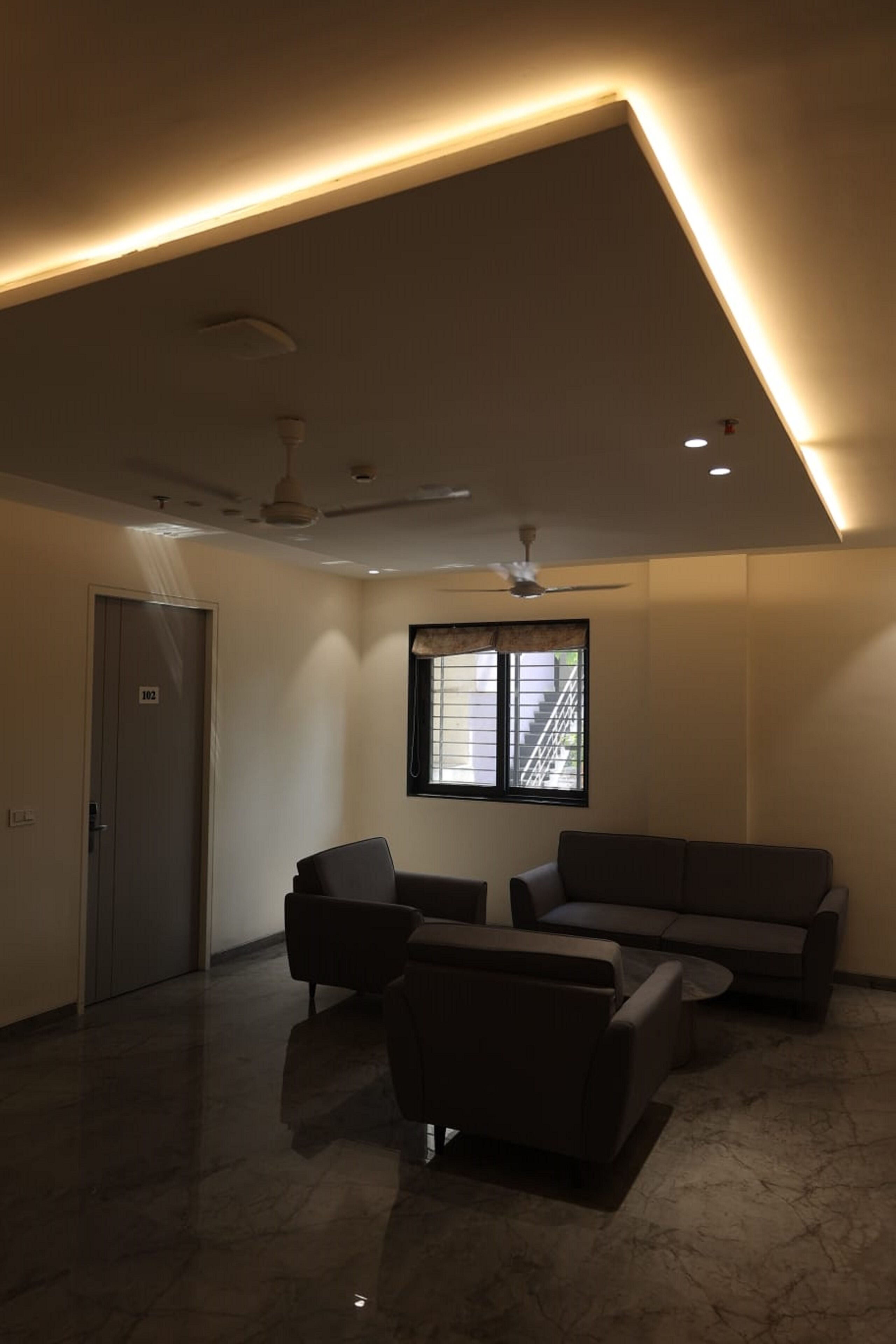 Un vaste espace de vie au design moderne, comprenant un grand canapé, un fauteuil plus petit et une table basse. On y trouve une fenêtre avec des stores, un ventilateur de plafond et un éclairage encastré.