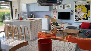 Una cocina moderna con una isla central, taburetes de bar, una mesa de comedor con una taza roja y una sala de estar con sofá y televisor.