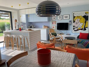 A modern kitchen with a central island, bar stools, a dining table with a red mug, and a living area with a sofa and a television. - In the heart of the countryside, close to the most beautiful sites in Périgord Noir. (Cénac-et-Saint-Julien)