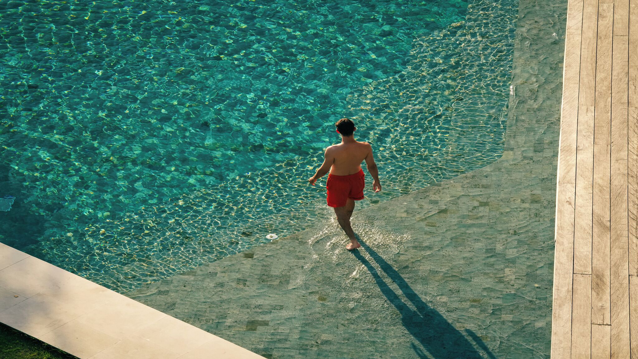 A man walking along the edge of a clear pool with a wooden deck.