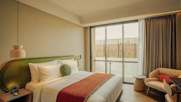 A hotel room with a large bed, a green headboard, a small table, a chair, and a view through a large window.