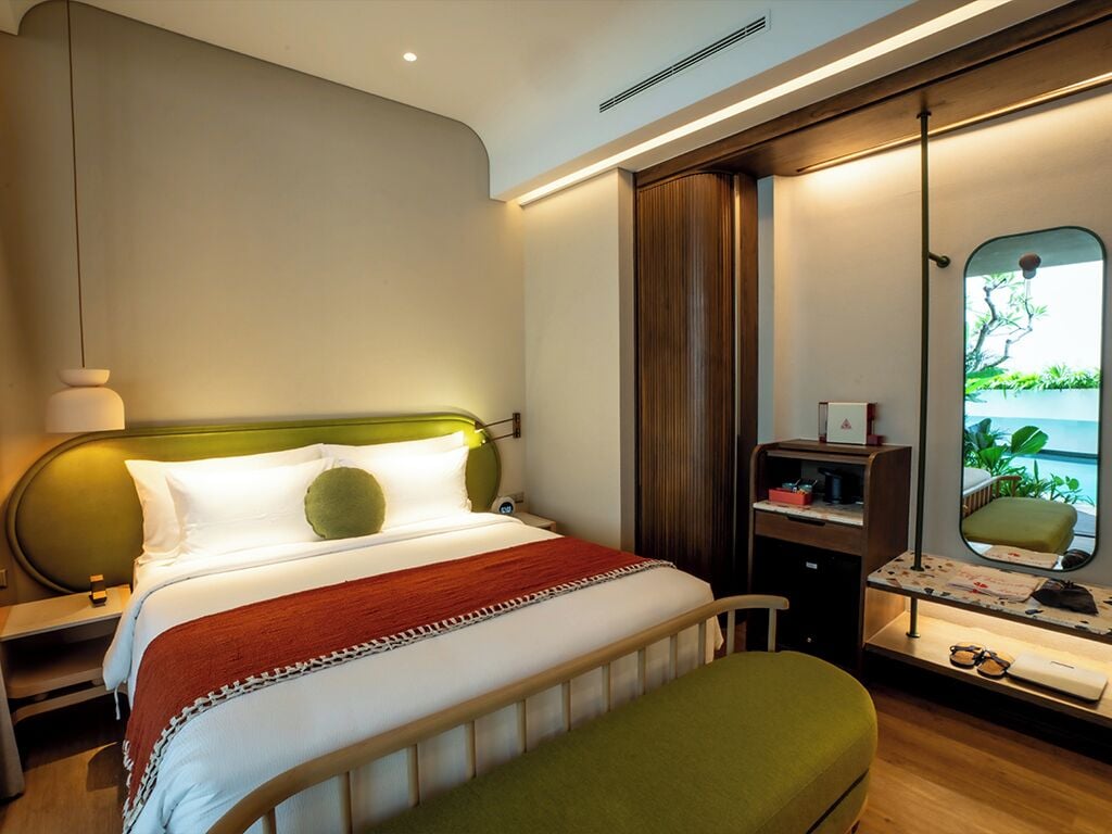 A modern hotel room with a bed, bedside table, nightstand, and a mirror with a view of greenery.