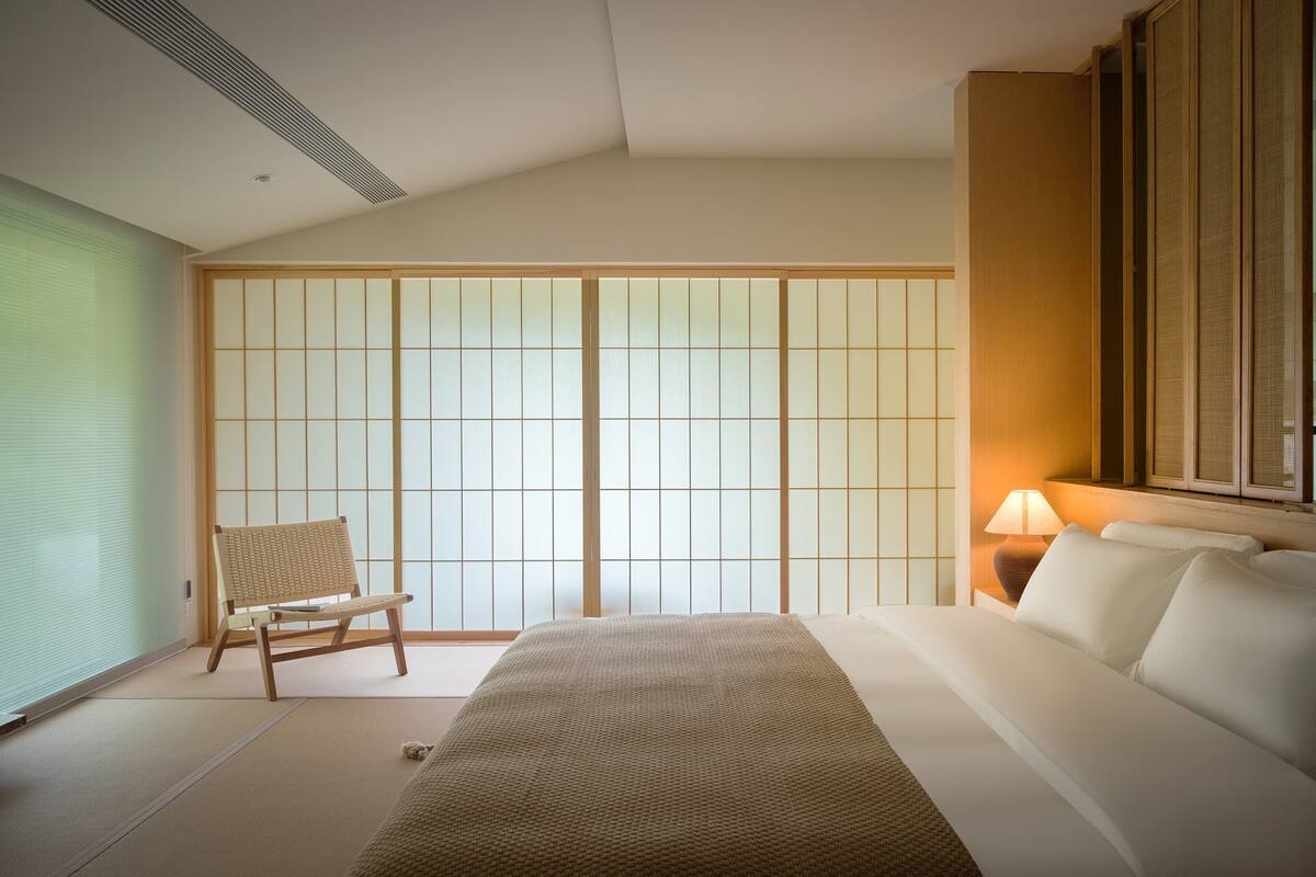 A traditional Japanese-style bedroom with tatami flooring, a low wooden bed, a shoji screen door, and a small wooden chair.