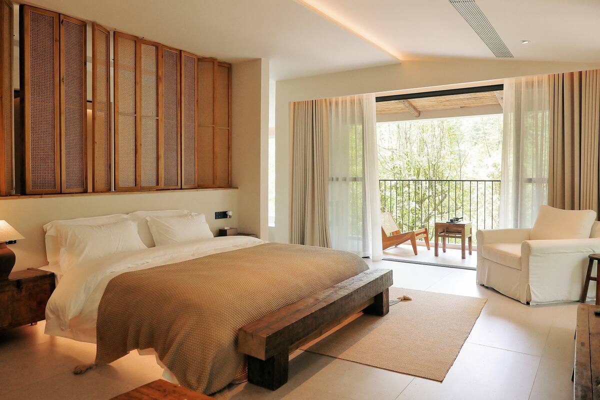 A spacious bedroom with a large bed, a wooden headboard, a balcony with a chair, and a view of greenery.