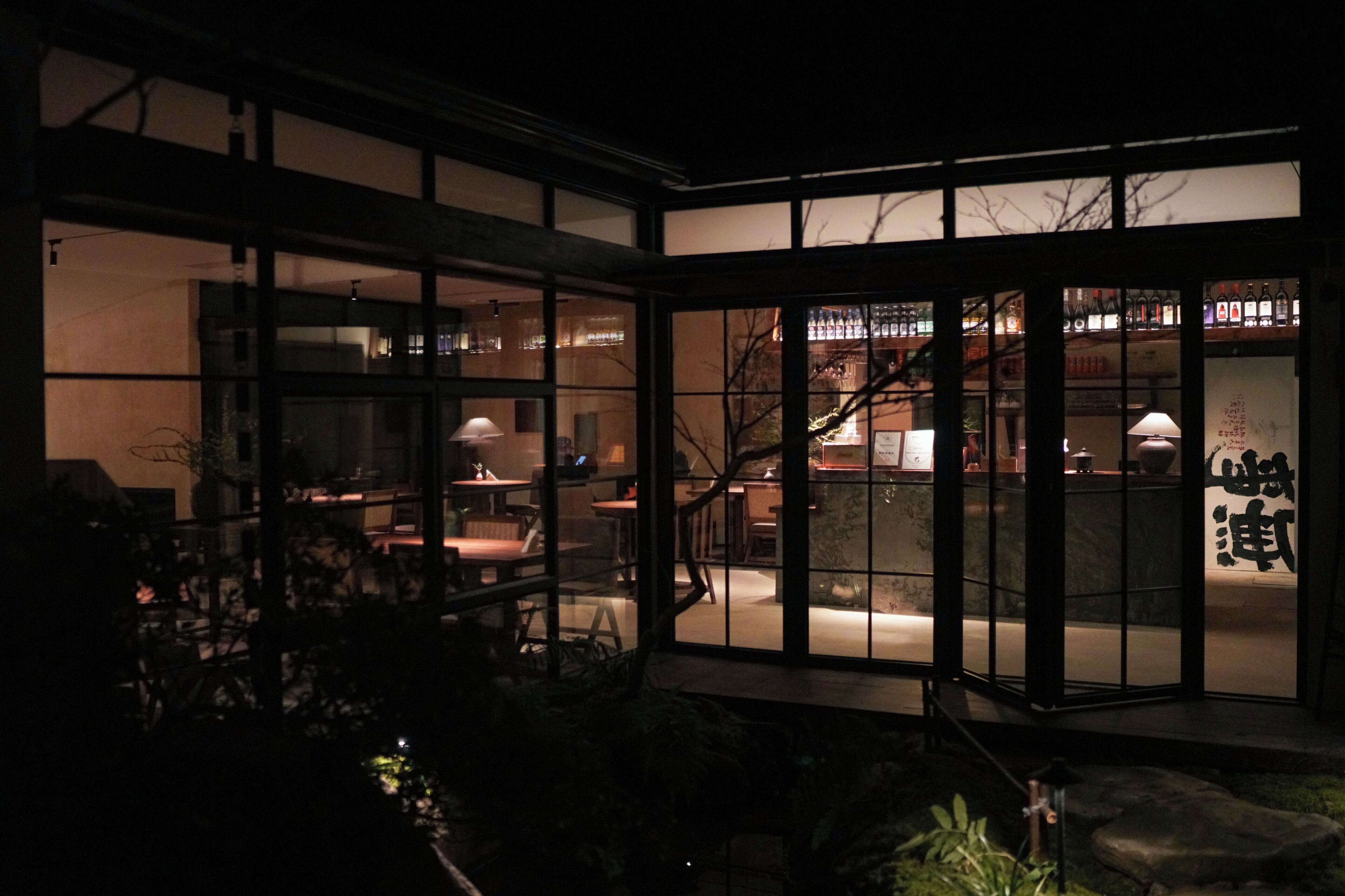 A dimly lit interior with a bar area visible through large windows, featuring a variety of bottles and a seating arrangement.