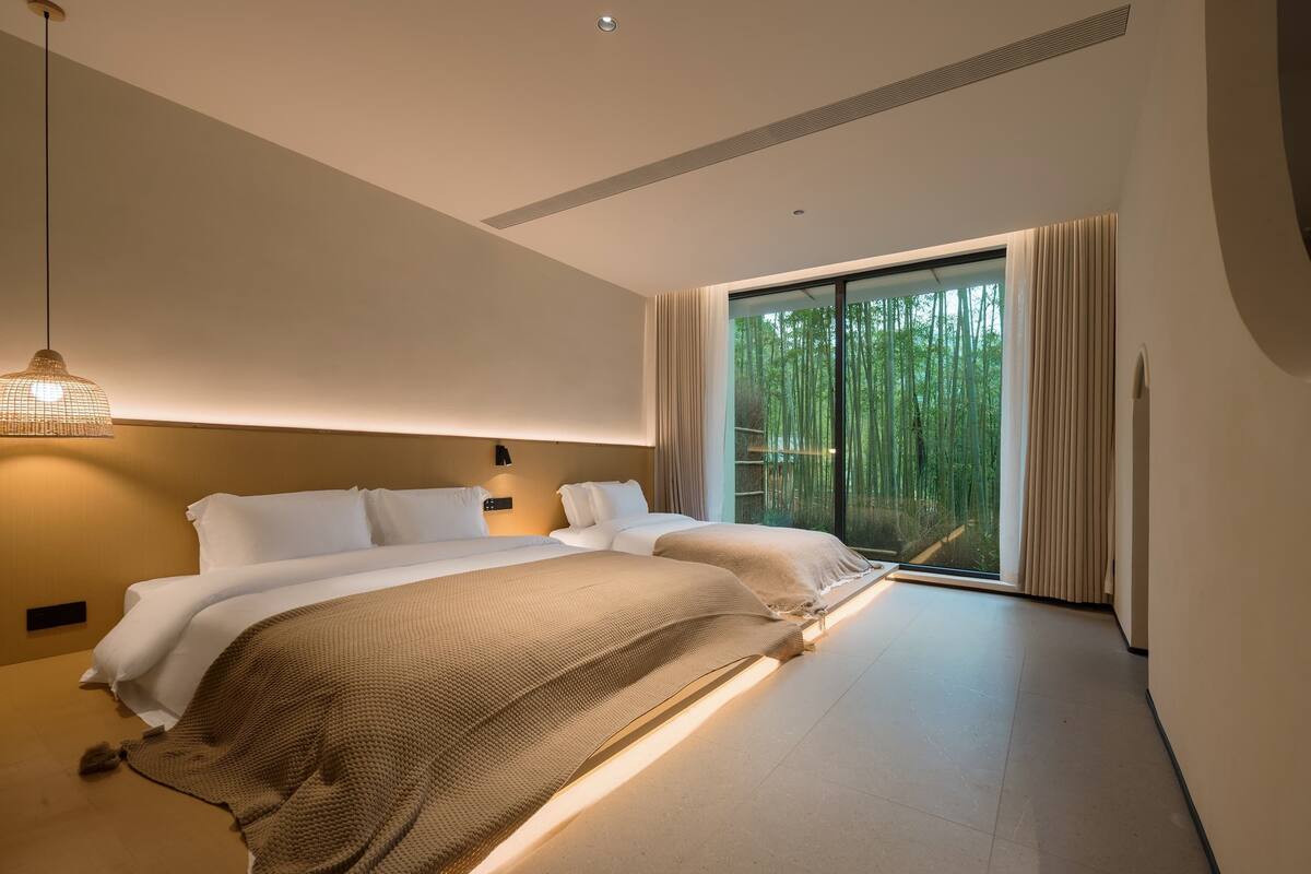 A modern bedroom with a large bed, a bedside lamp, and a view of bamboo trees through a sliding glass door.