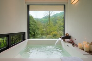 A modern bathroom with a large bathtub, a window with a forest view, and candles on the side. - BuSu Forest Forever Hotel (Chengdu)
