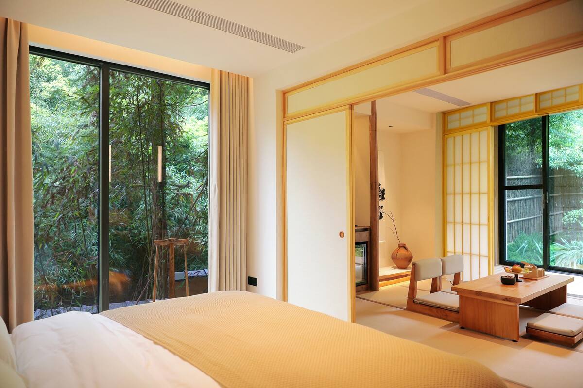A modern hotel room with a large sliding glass door leading to a balcony, a bed with a beige bedspread, a wooden desk with a chair, and traditional Japanese sliding doors.