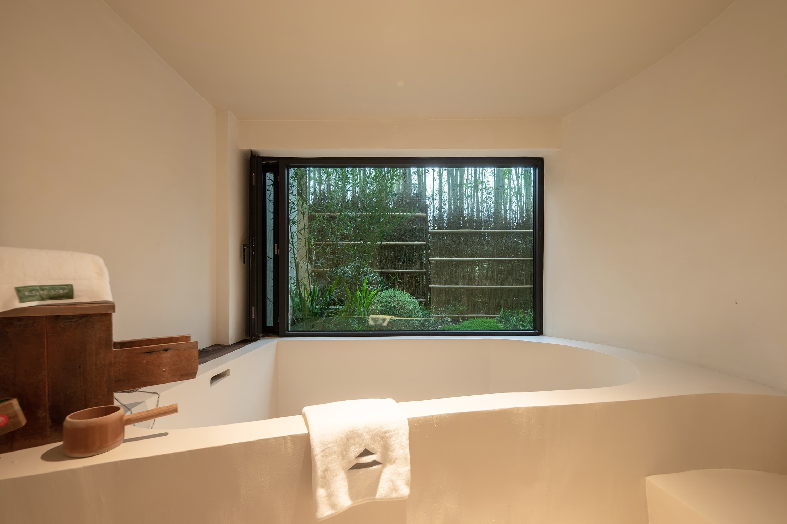 Un baño moderno con una bañera grande, una mesita de madera y una ventana con vistas a un jardín de bambú.