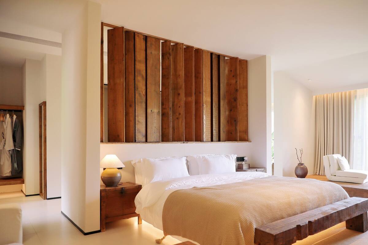 A modern bedroom with a large bed, wooden headboard, bedside tables, a bench, and a view of a cityscape through the window.