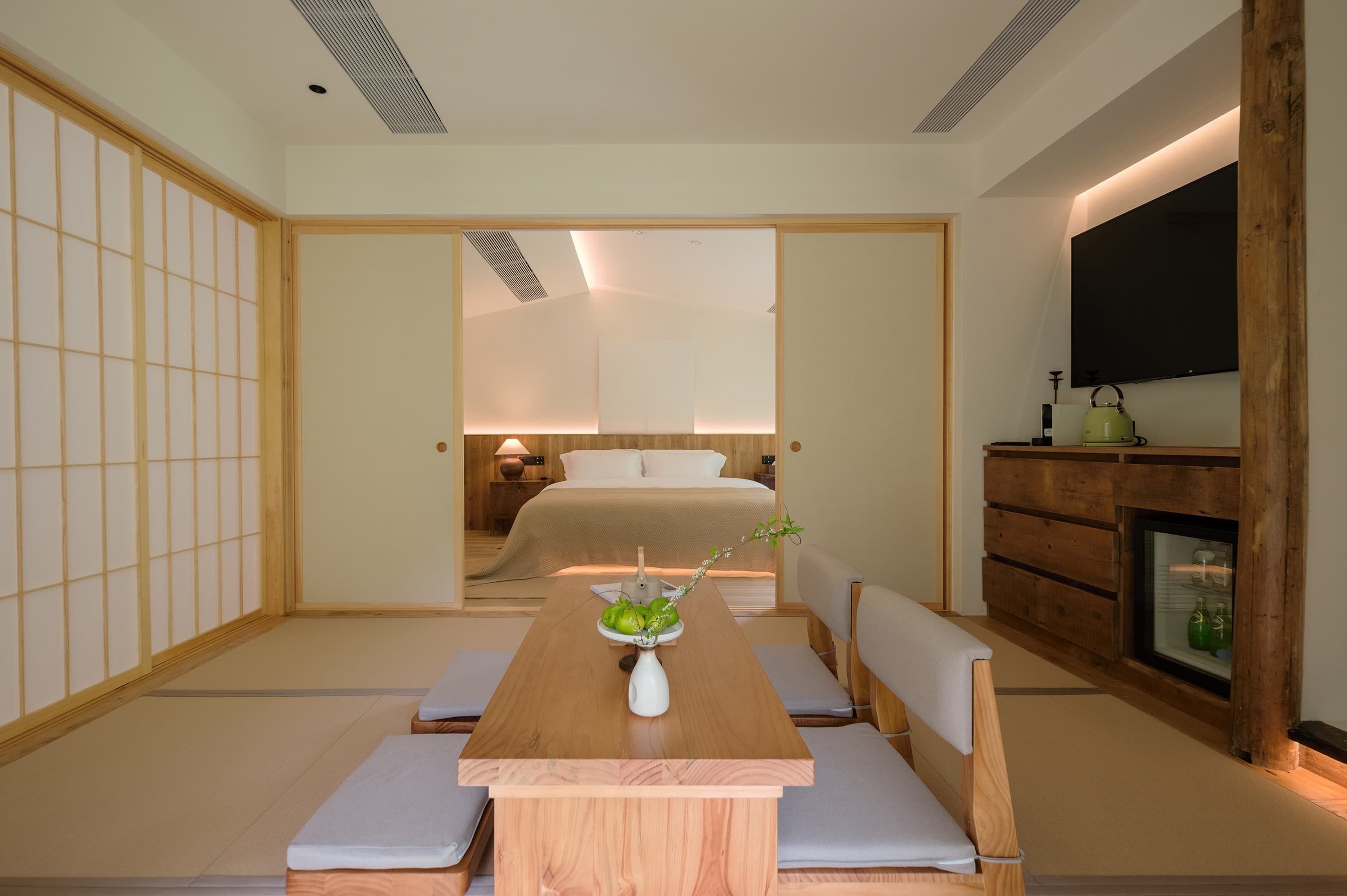 A traditional Japanese-style room with tatami flooring, a low wooden table with cushions, a bed with a white headboard, and a television mounted on the wall.
