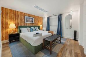A modern bedroom with a wooden wall panel, a green bed, two bedside tables with lamps, a large mirror, and a window with blue curtains. - 6k sq/ft Mansion! Heated Pool, Hot Tub, Game Room, Theatre, Unforgettable Views! (Colorado Springs)