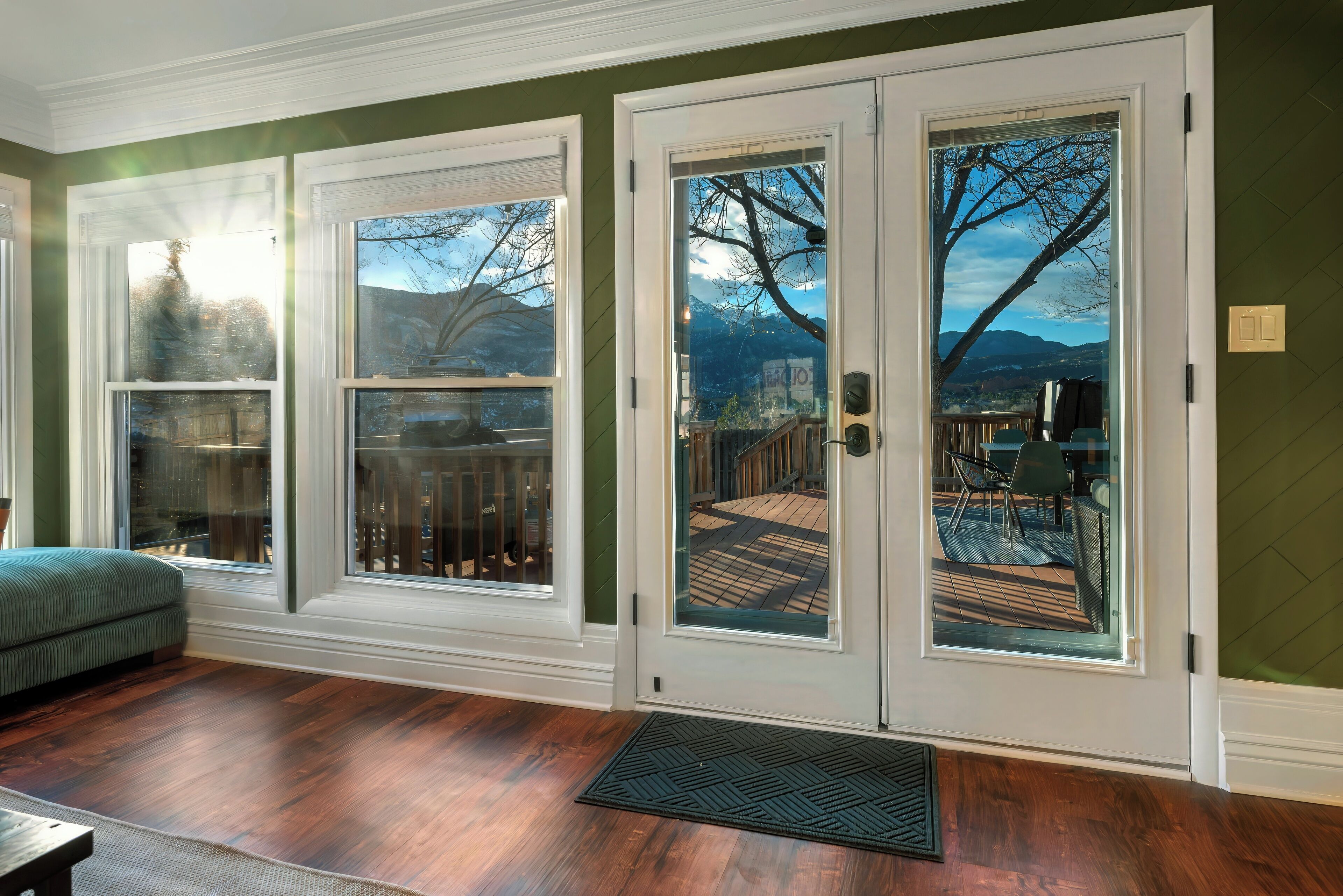 A room with a wooden floor, a green couch, and a view of a deck and mountains through glass doors.