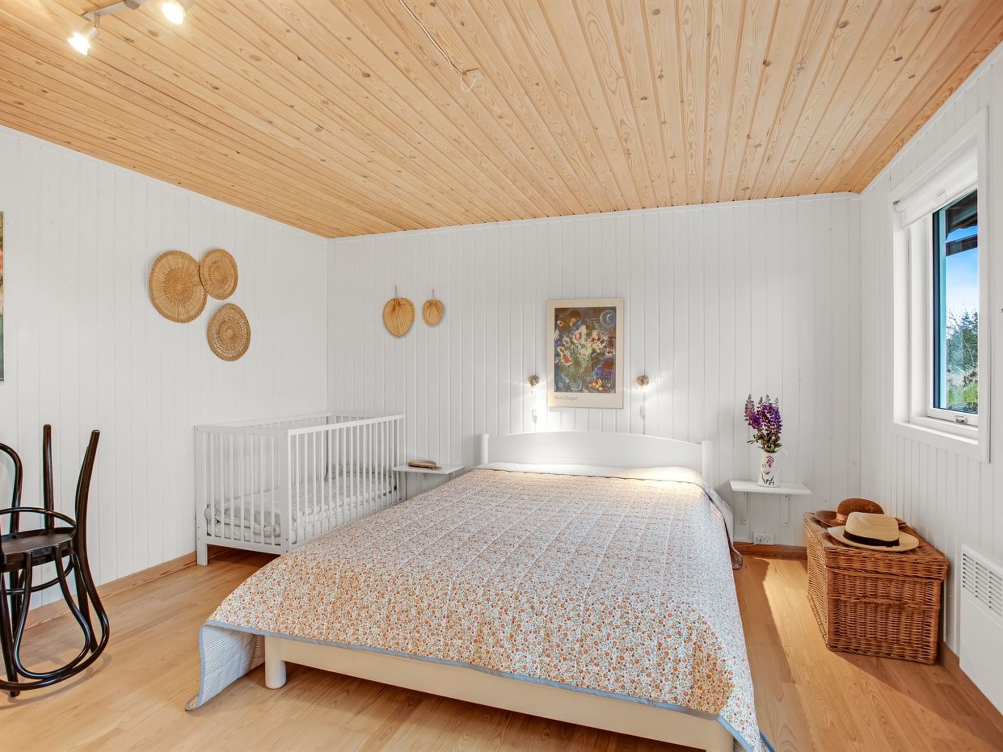 A bedroom with a wooden ceiling, white walls, a bed with a floral bedspread, a white crib, a wicker basket, and a window with a view.