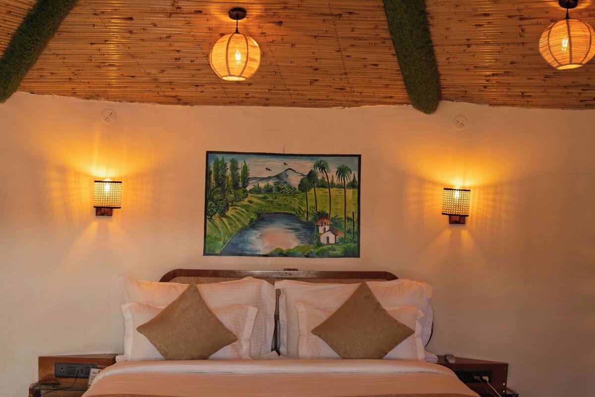 A bedroom with a wooden ceiling, a large bed with white bedding, two wall-mounted lamps, and a painting of a landscape on the wall.